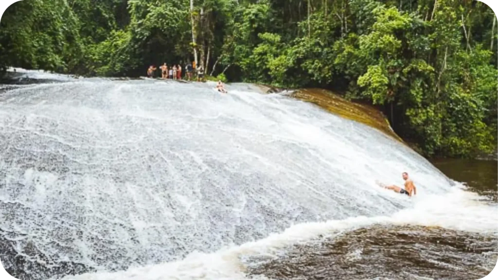 Cachoeira com grande pedra inclinada por onde pessoas escorregam como em um tobogã natural, rodeada por vegetação da mata atlântica, em Paraty, RJ