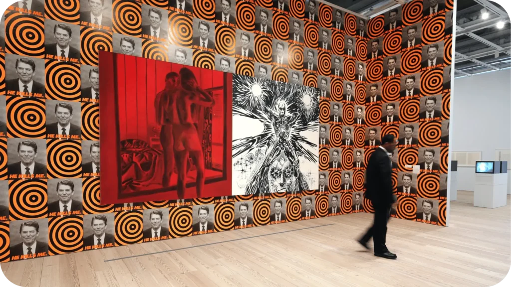 "Homem caminha por galeria de arte contemporânea com paredes cobertas por imagens repetidas de Ronald Reagan com alvos laranja e a frase 'HE KILLS ME'. No centro da parede, duas obras em destaque: uma pintura vermelha de figuras nuas de costas e uma arte gráfica em preto e branco com figura humana estilizada em meio a explosões visuais."