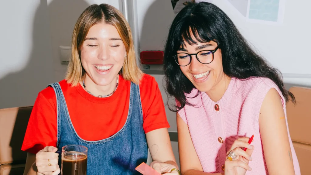 Duas mulheres sorridentes em um ambiente de trabalho colaborativo, compartilhando um momento de alegria enquanto seguram uma caneta e uma bebida, representando a leveza e os laços positivos promovidos pela felicidade corporativa.