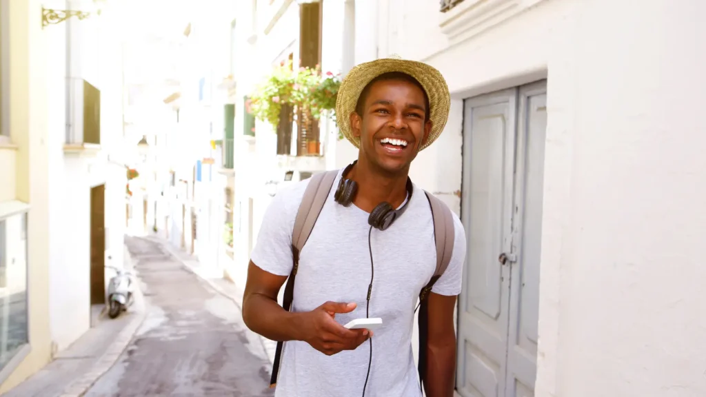Homem jovem sorridente caminhando por uma rua urbana clara, usando chapéu, mochila e fones de ouvido enquanto olha o celular, imagem que remete a um estilo de vida saudável e leve frequentemente explorado em campanhas de wellness washing sem ações reais de bem-estar.