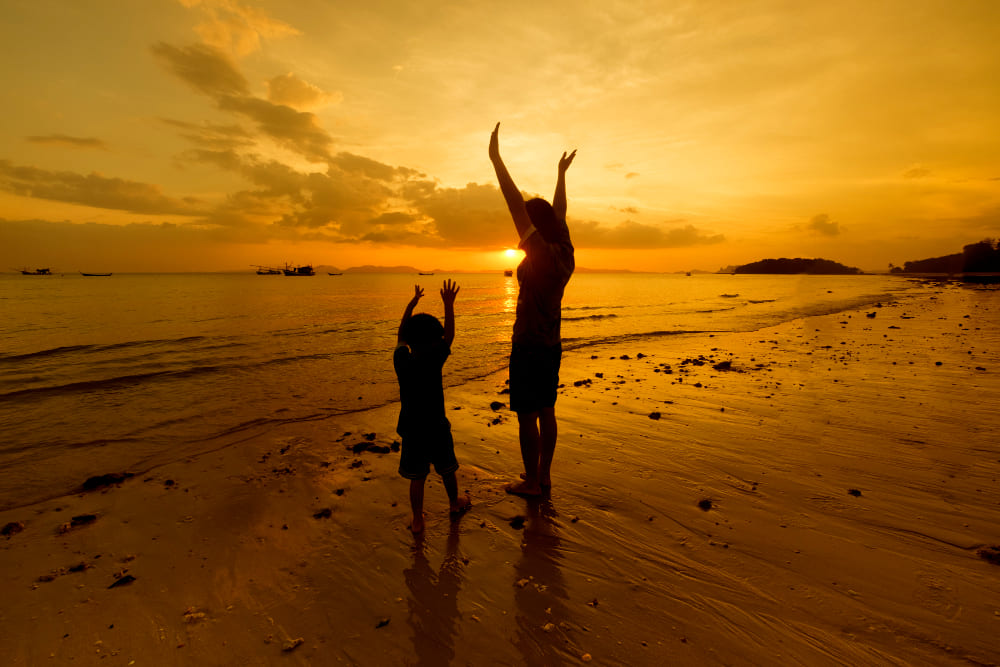 silhueta de adulto e criança com braços erguidos na praia ao pôr do sol celebrando benefício de viagem corporativo.