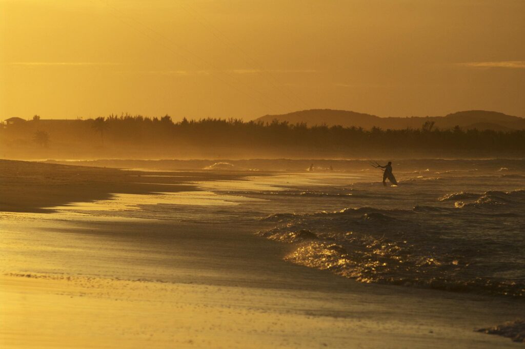 silhueta de kitesurfista ao pôr do sol dourado em praia do nordeste em julho com ventos ideais para o esporte.