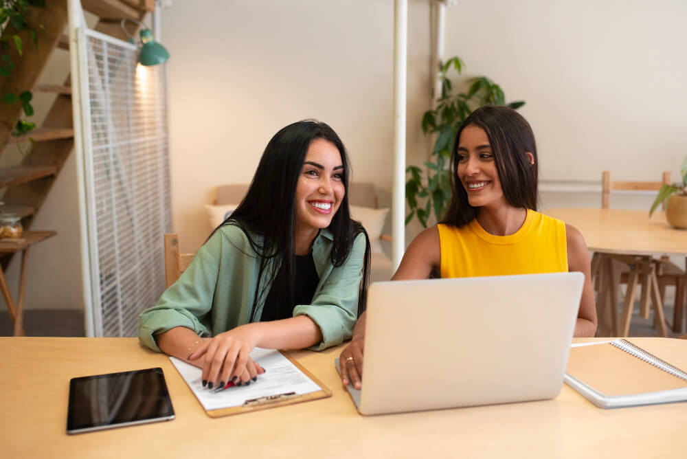 duas colaboradoras sorrindo juntas diante de notebook em ambiente de trabalho positivo e acolhedor.
